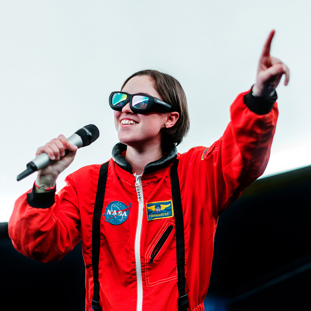 Tjej klädd som astronaut håller i en mikrofon och pekar mot publiken.