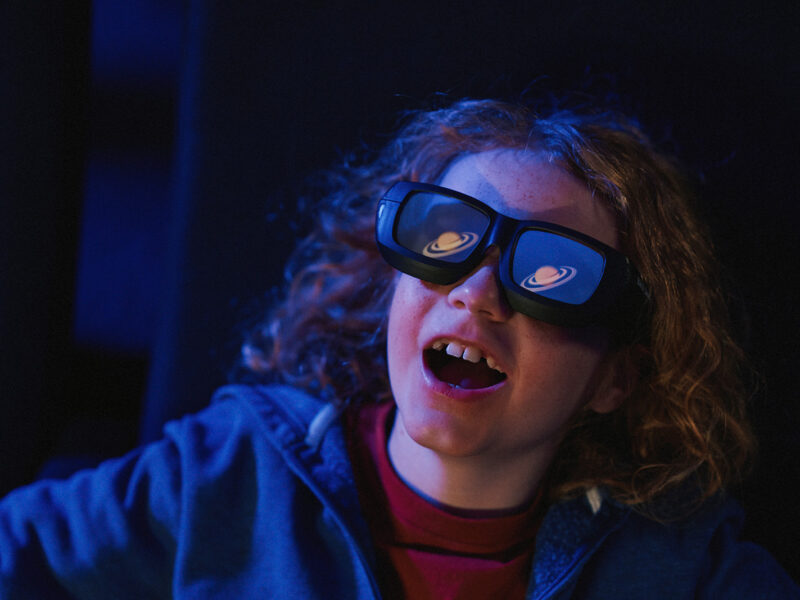 ung tjej med lockigt hår som tittar häpet mnedan hennes 3D-glasögon reflekterar en planet.